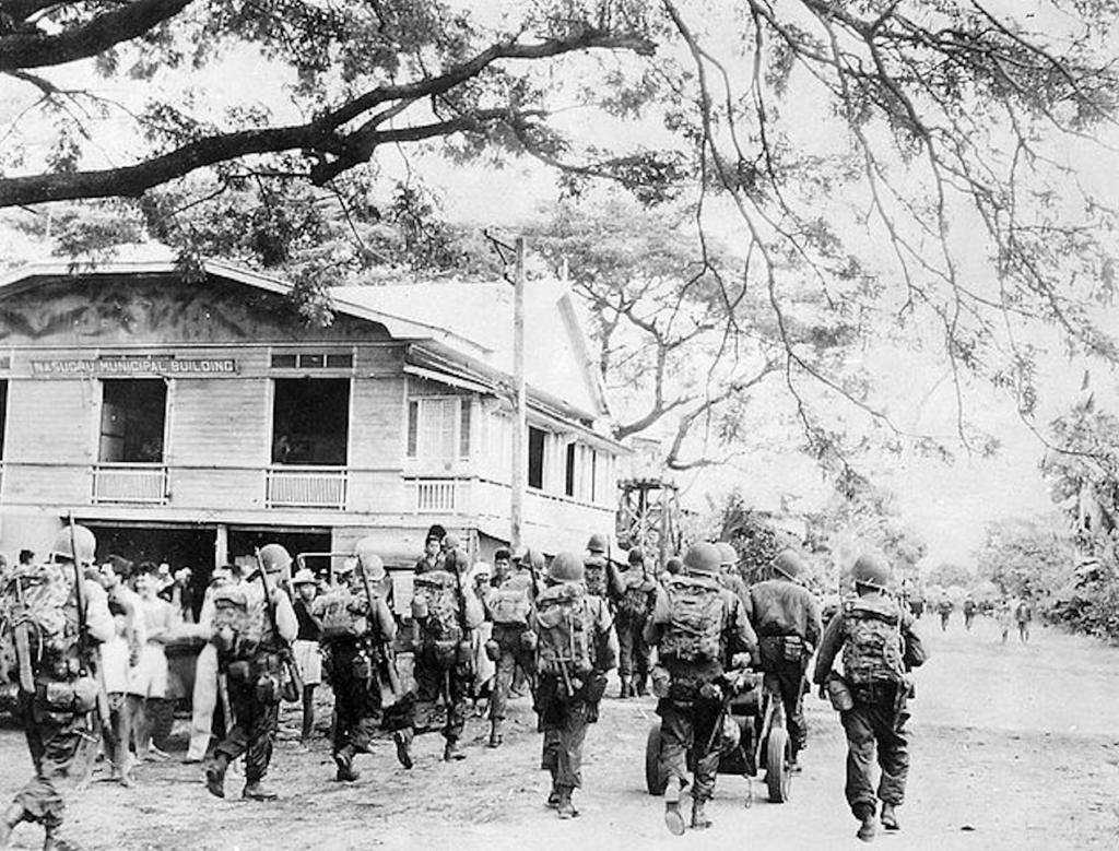 Troops of the 188th Glider Infantry Regiment, 11th Airborne Division, make their way through the town of Nasugbu on the island of Luzon, 31 January 1945.