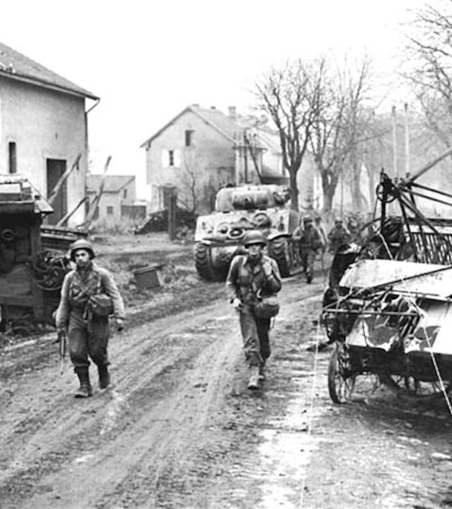 Black and white photo of soldiers walking down the unpaved road in the town of Metz. Abandoned vehicles lie along the road side.